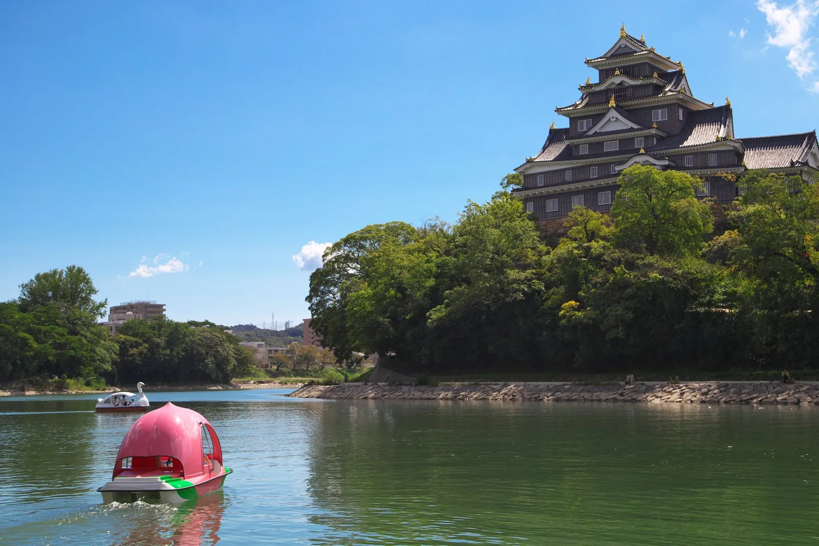 旭川に浮かぶ桃の形のボートと、木々の向こうに建つ黒い岡山城